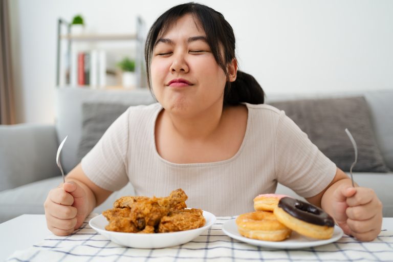 居家餐桌前，一名女性面對炸雞與甜甜圈等高熱量食物，準備進食的情境，呈現肥胖與飲食習慣的關聯主題。